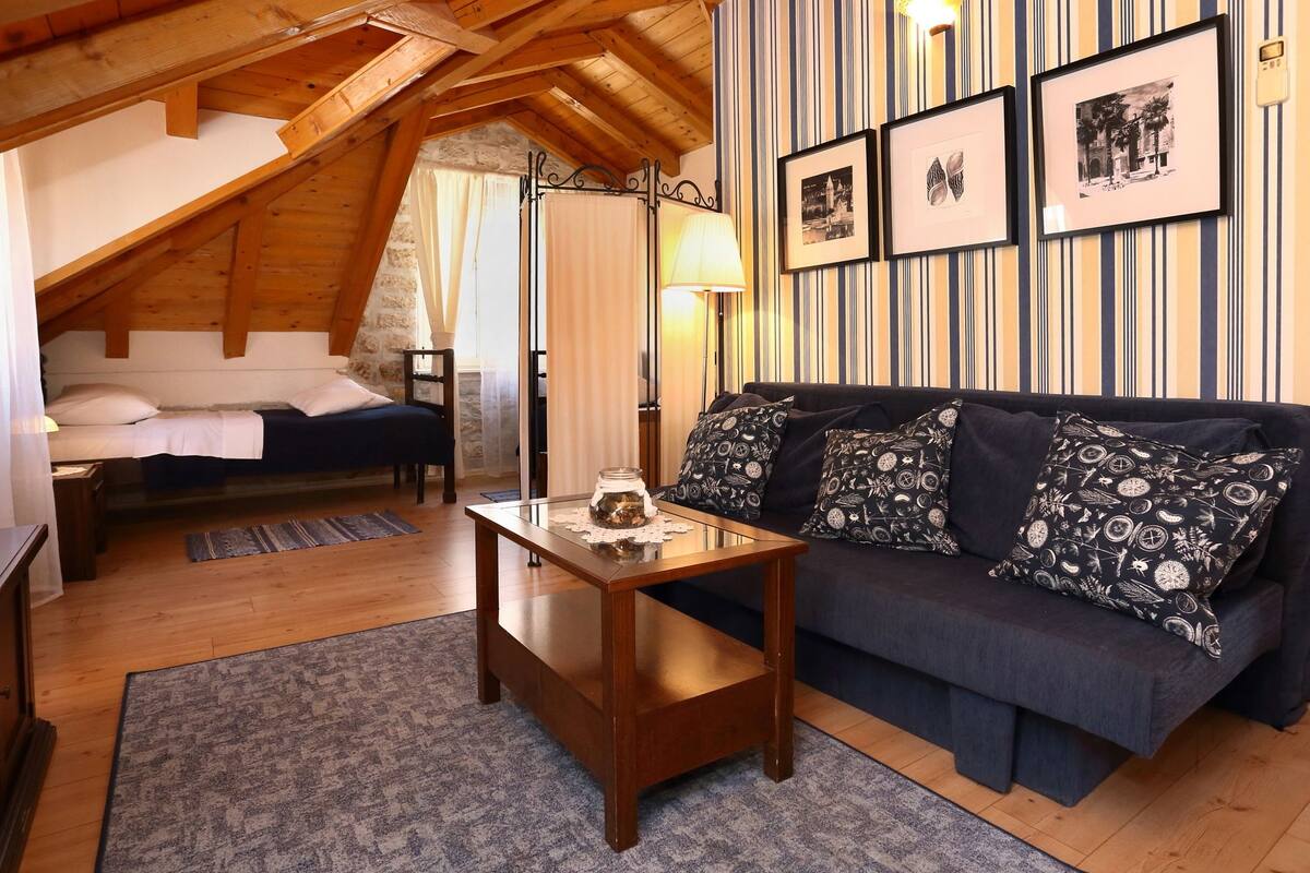 A cozy attic bedroom with a wooden ceiling, a dark blue sofa with patterned cushions, a small wooden table, and a bed with white linens.