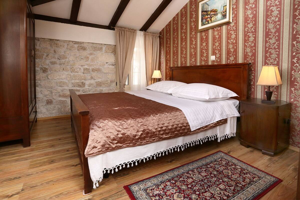 A bedroom with a wooden bed, a stone wall, a patterned wallpaper, and a red and beige rug.