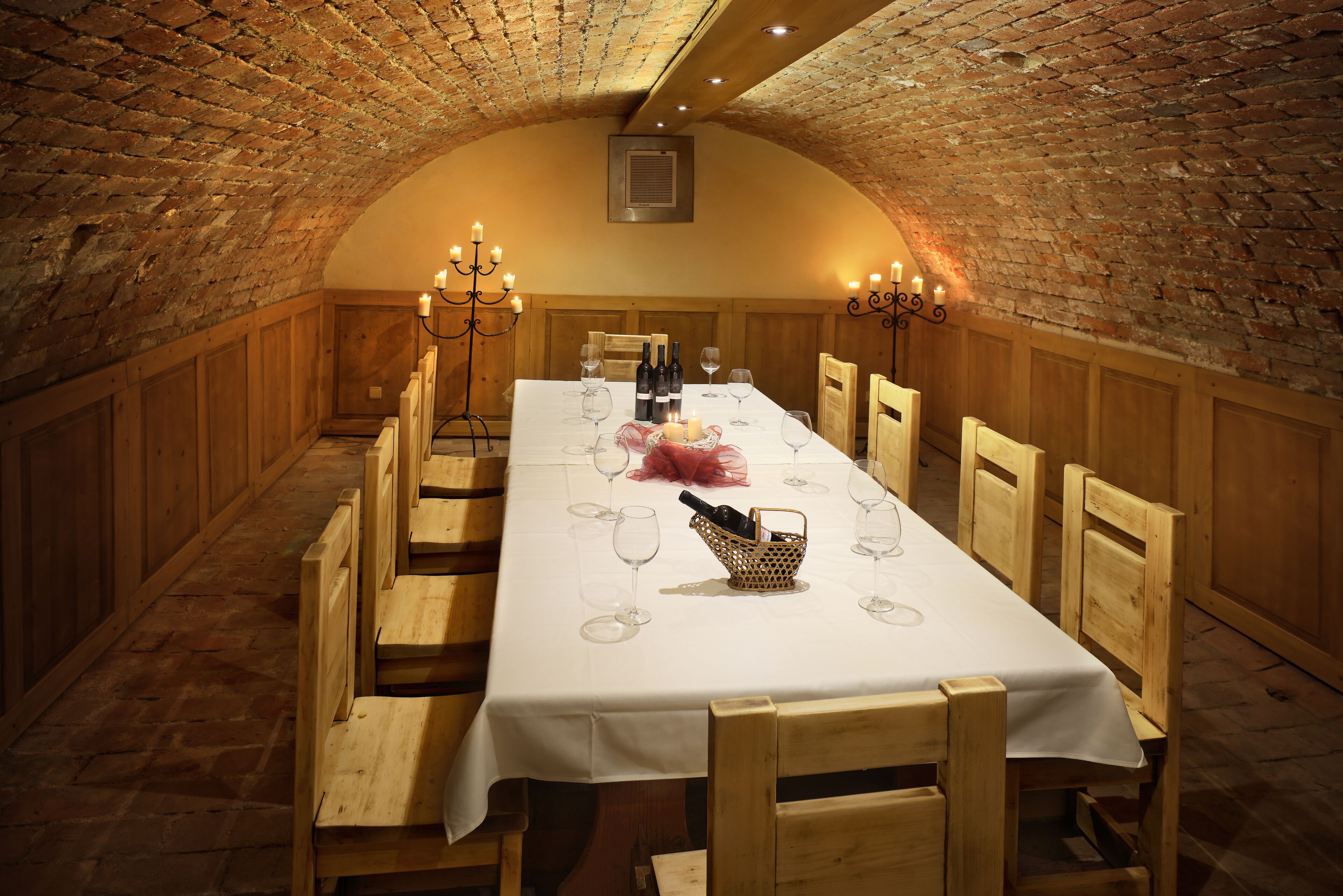 A long dining table set with wine glasses, bottles, and a candle centerpiece in a brick-walled room with wooden benches.