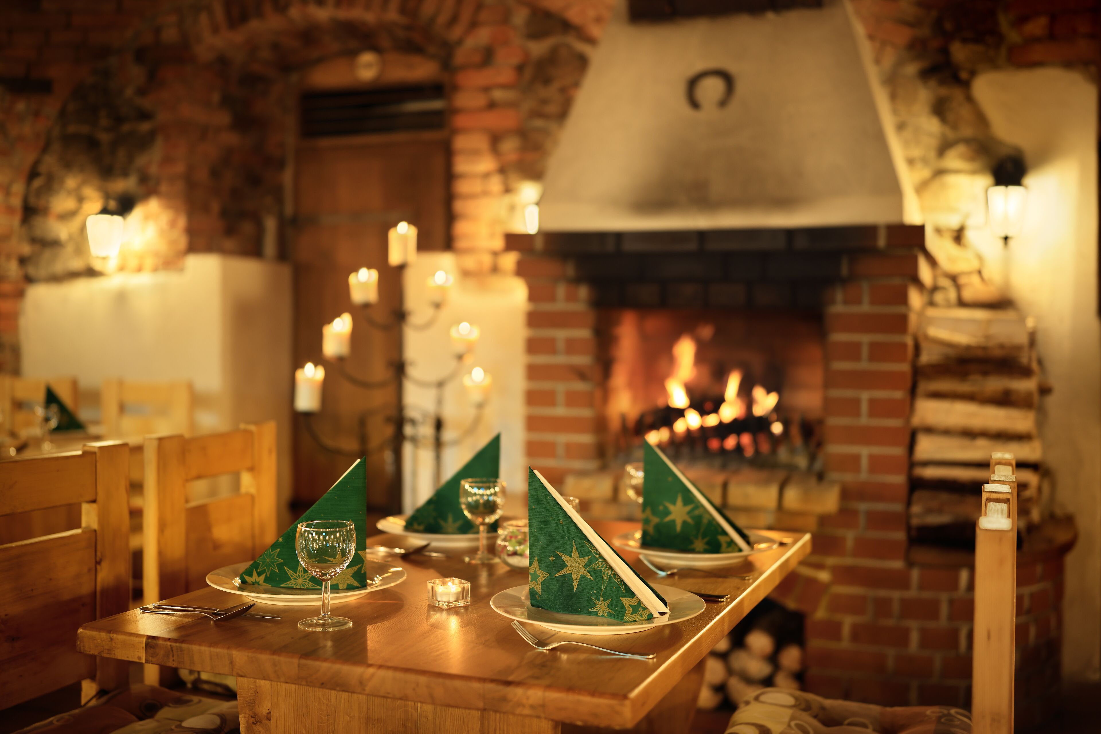 Mesa de comedor dispuesta con servilletas verdes, copas de vino y velas, con una chimenea de ladrillo al fondo.