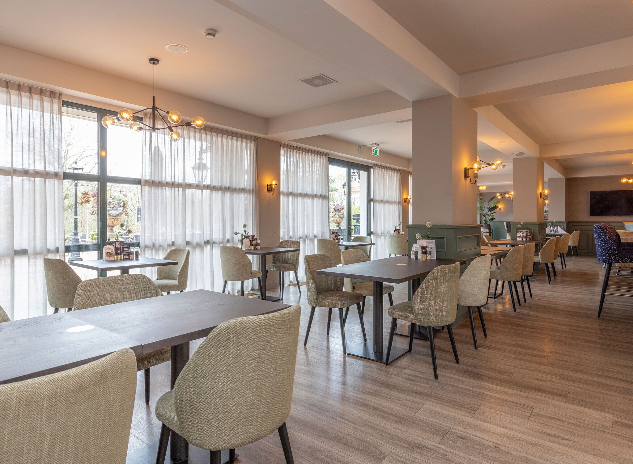 A spacious dining area with multiple tables and chairs, large windows with curtains, and a modern chandelier.