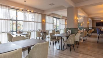 A spacious dining area with multiple tables and chairs, large windows with curtains, and a modern chandelier.