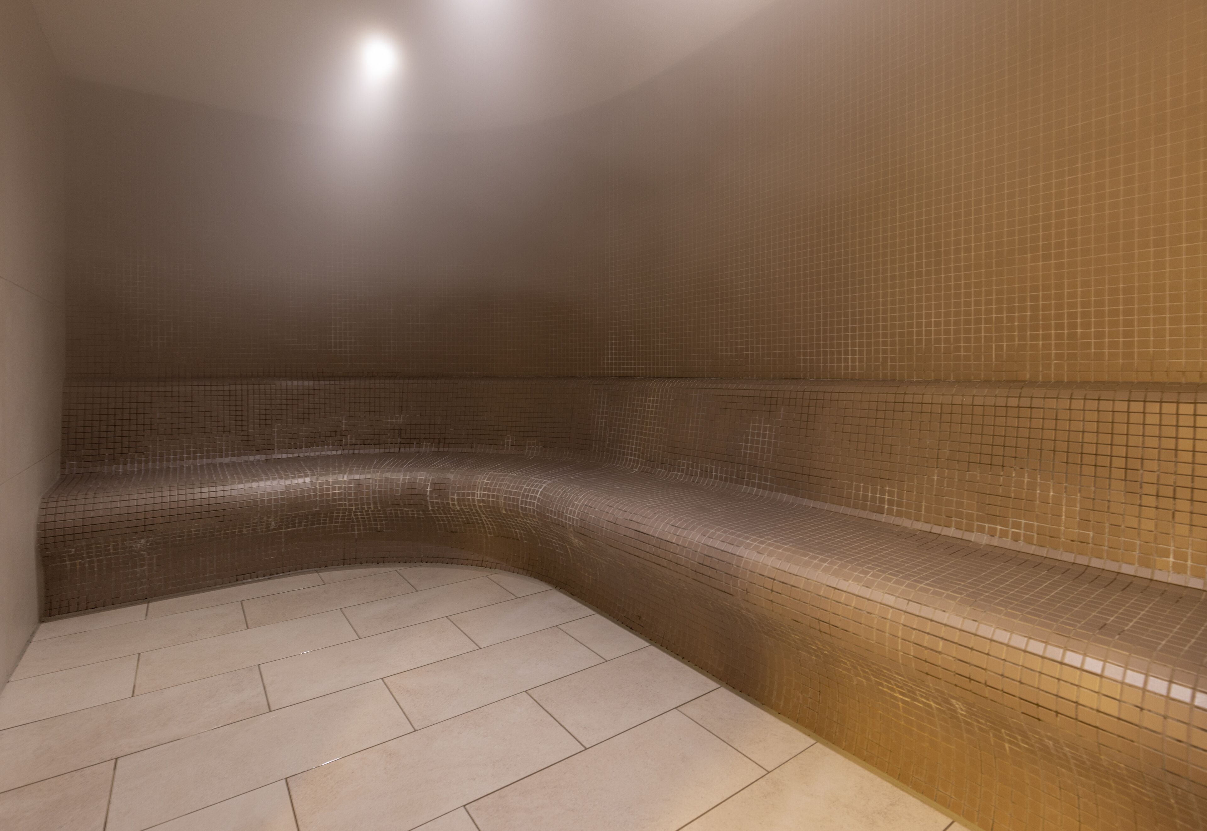 A steam room with tiled walls and floor, a bench, and a light fixture.
