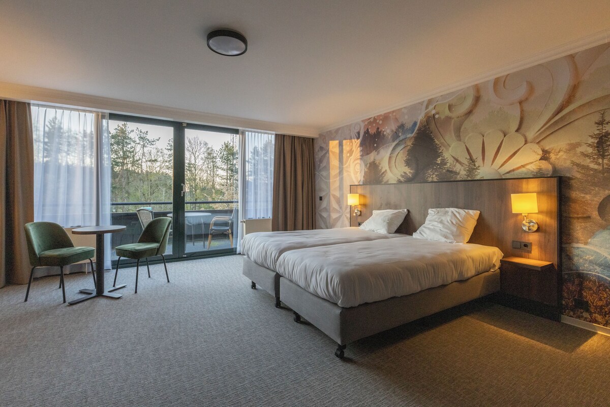 Hotelkamer met een groot bed, twee nachtkastjes met lampen, een kleine tafel met twee stoelen, en uitzicht op bomen door grote ramen.