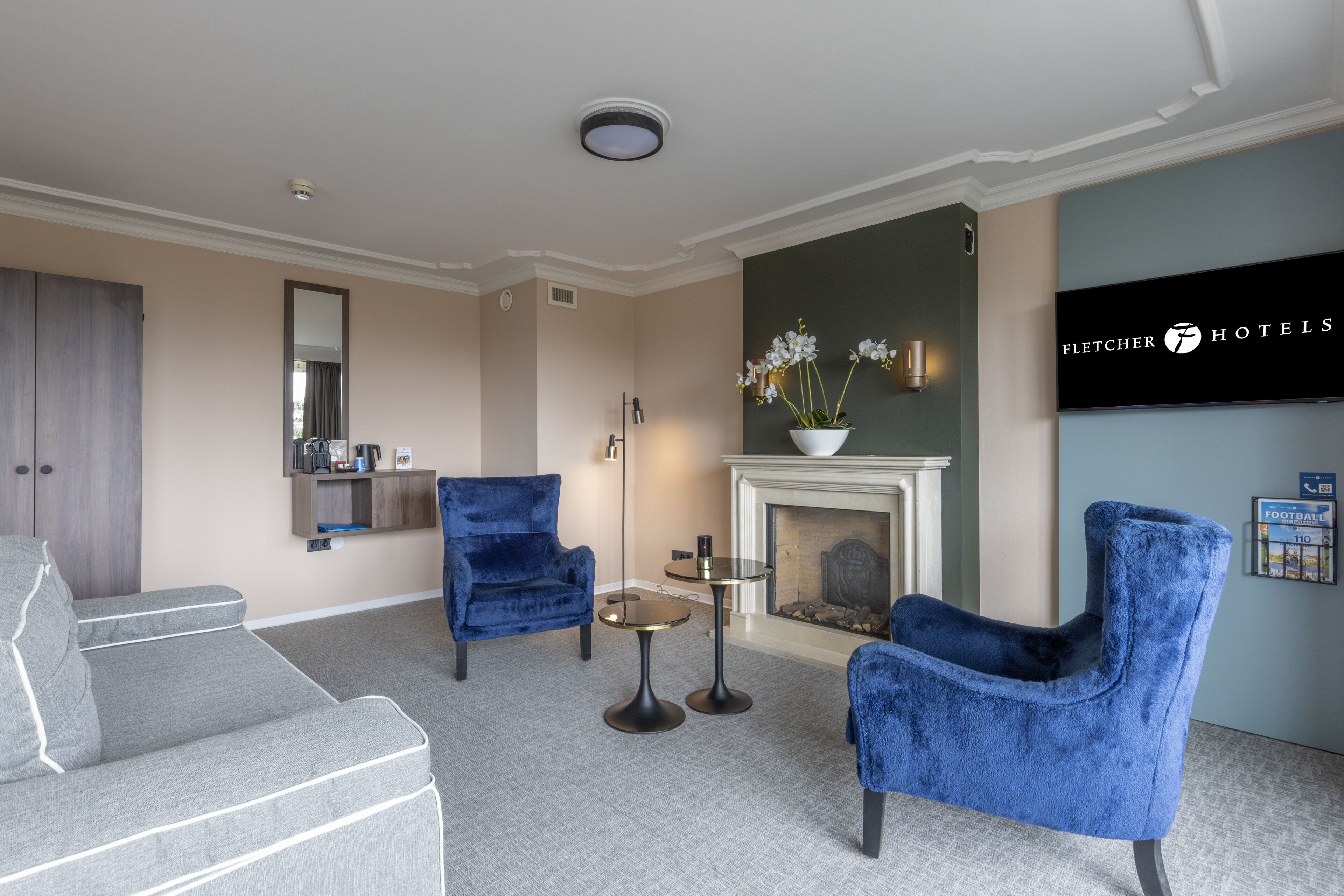 A hotel lobby with a fireplace, two blue armchairs, a grey sofa, a small round table, and a mirror.
