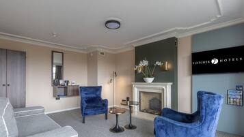 A hotel lobby with a fireplace, two blue armchairs, a grey sofa, a small round table, and a mirror.