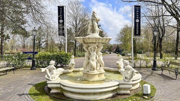 A classical fountain with statues, surrounded by a paved area with benches and banners for hotels.