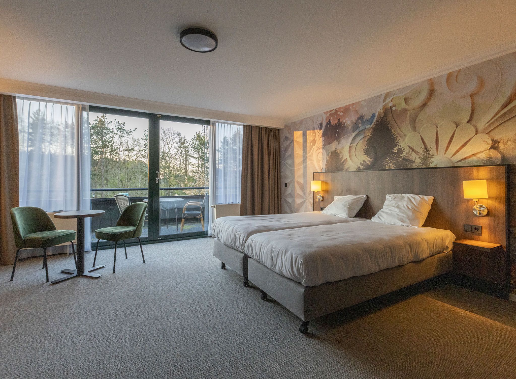 A hotel room with a large bed, a chair, a table, and a view of trees through the window.