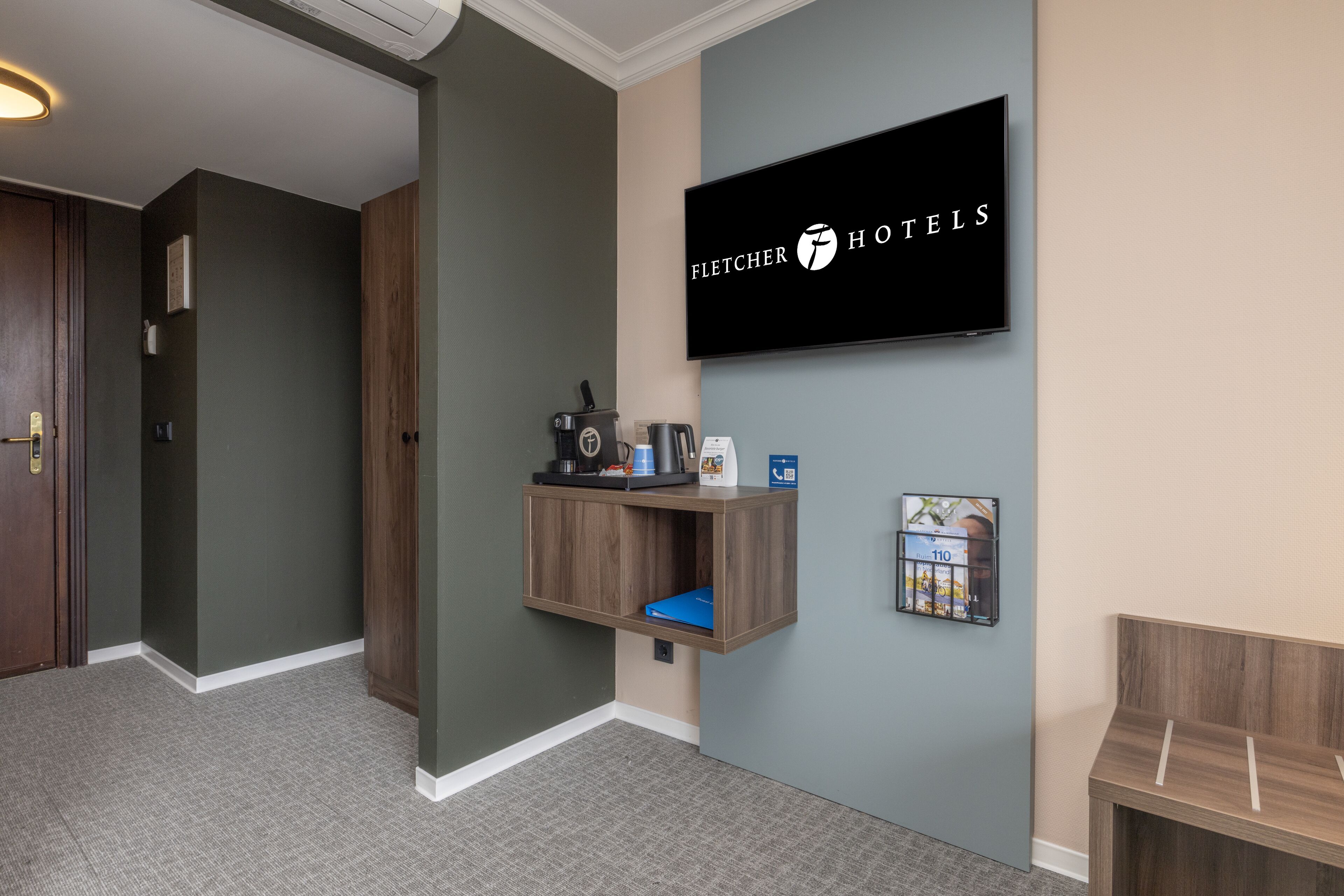 Zona reception di un hotel con una televisione a parete che mostra il logo 'Fletcher Hotels', uno scaffale in legno con una macchina per il caffè, un telefono e un quadro incorniciato.