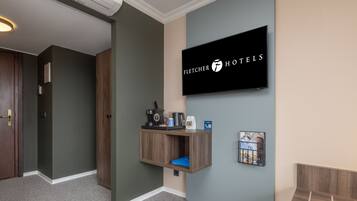 Zona reception di un hotel con una televisione a parete che mostra il logo 'Fletcher Hotels', uno scaffale in legno con una macchina per il caffè, un telefono e un quadro incorniciato.