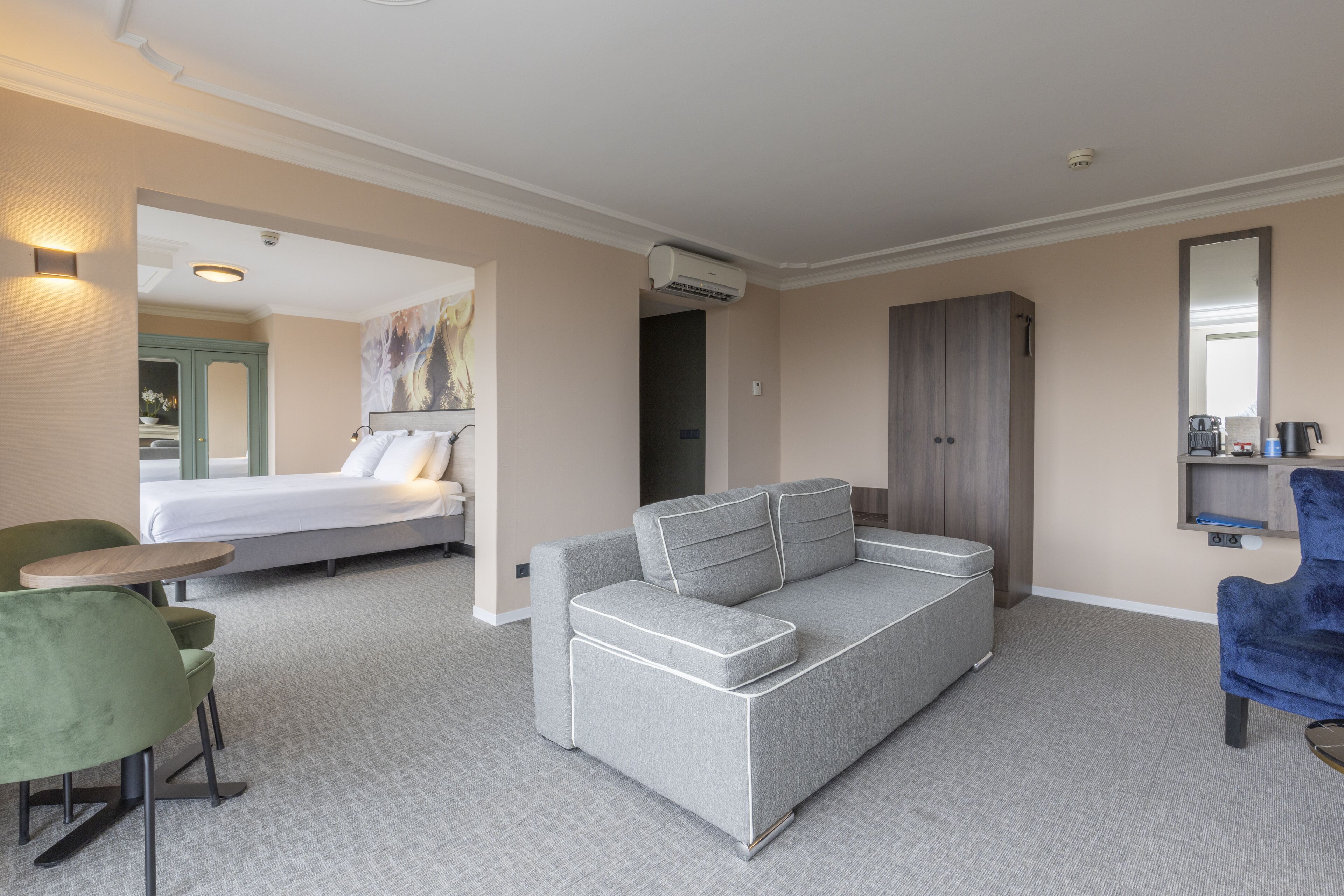 Una habitación de hotel moderna con un sofá gris, un sillón verde, una cama y un armario.