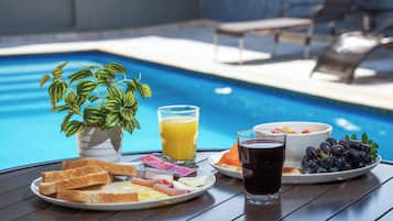 A breakfast setup by a pool with toast, eggs, ham, juice, and a bowl of fruit.