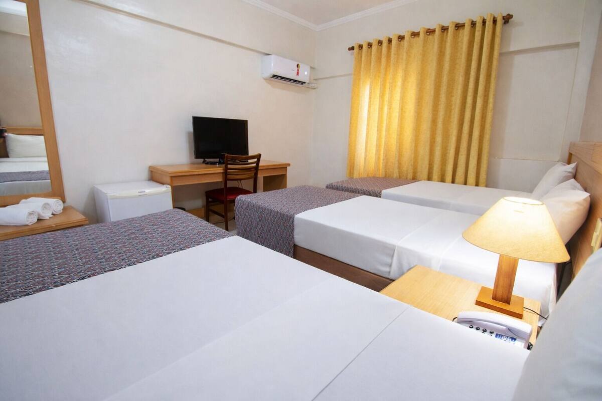 Ein Hotelzimmer mit drei Betten, einem Schreibtisch mit Stuhl, einem Fernseher und einer Lampe.