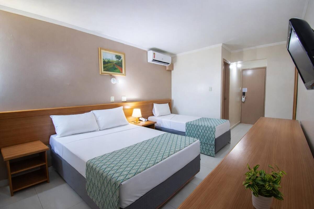 Ein Hotelzimmer mit zwei Betten, einem Schreibtisch, einem Fernseher, einem Gemälde und einer Klimaanlage.