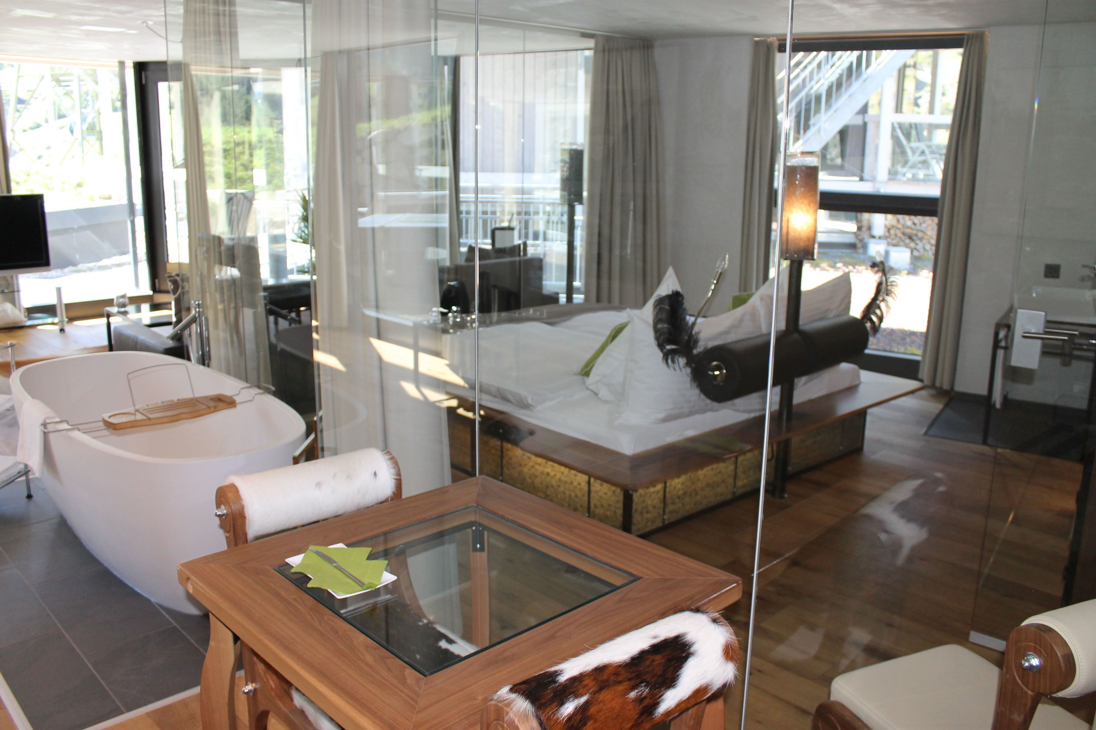 A modern hotel room with a large bed, a glass-enclosed bathroom, and a dining area with a table and chairs.
