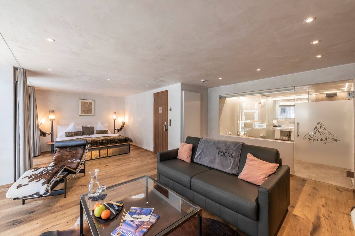 Une chambre d’hôtel moderne équipée d’un canapé, d’un fauteuil de détente, d’une table basse en verre avec des fruits et des magazines, d’un lit avec une tête de lit en bois et d’une kitchenette visible à travers une paroi en verre.