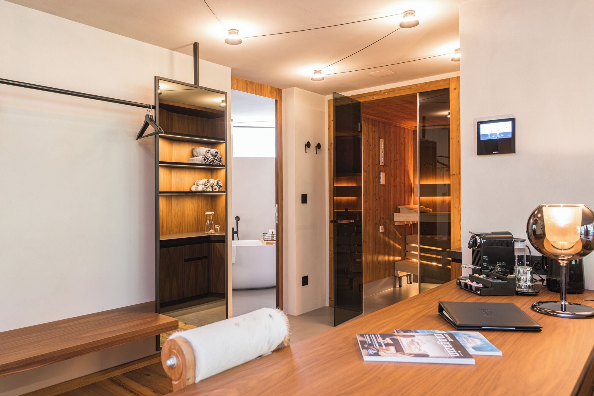 A modern hotel room with a wooden desk, a laptop, a coffee machine, and a large mirror.