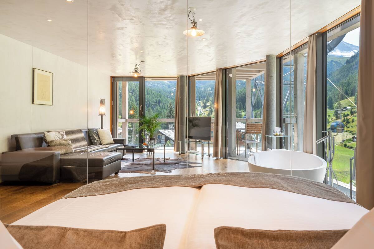 Une chambre spacieuse avec un grand lit, un coin salon avec un canapé et une table basse, une baignoire et un balcon offrant une vue sur la montagne.