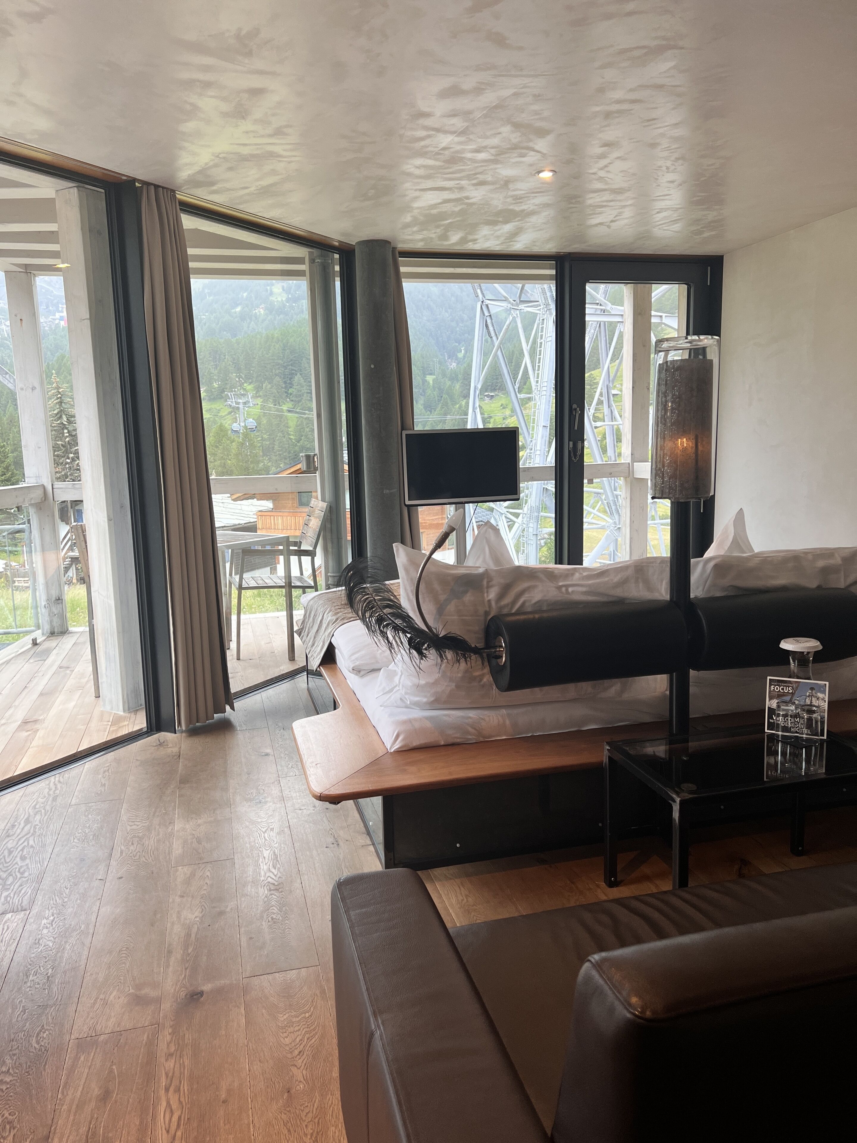 A modern hotel room with a large bed, a wooden bench, a glass coffee table, and a view of a mountain and cable car.