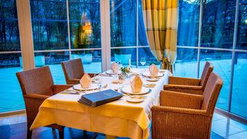 A dining area with a table set for four, surrounded by wicker chairs, large windows, and a view of trees.