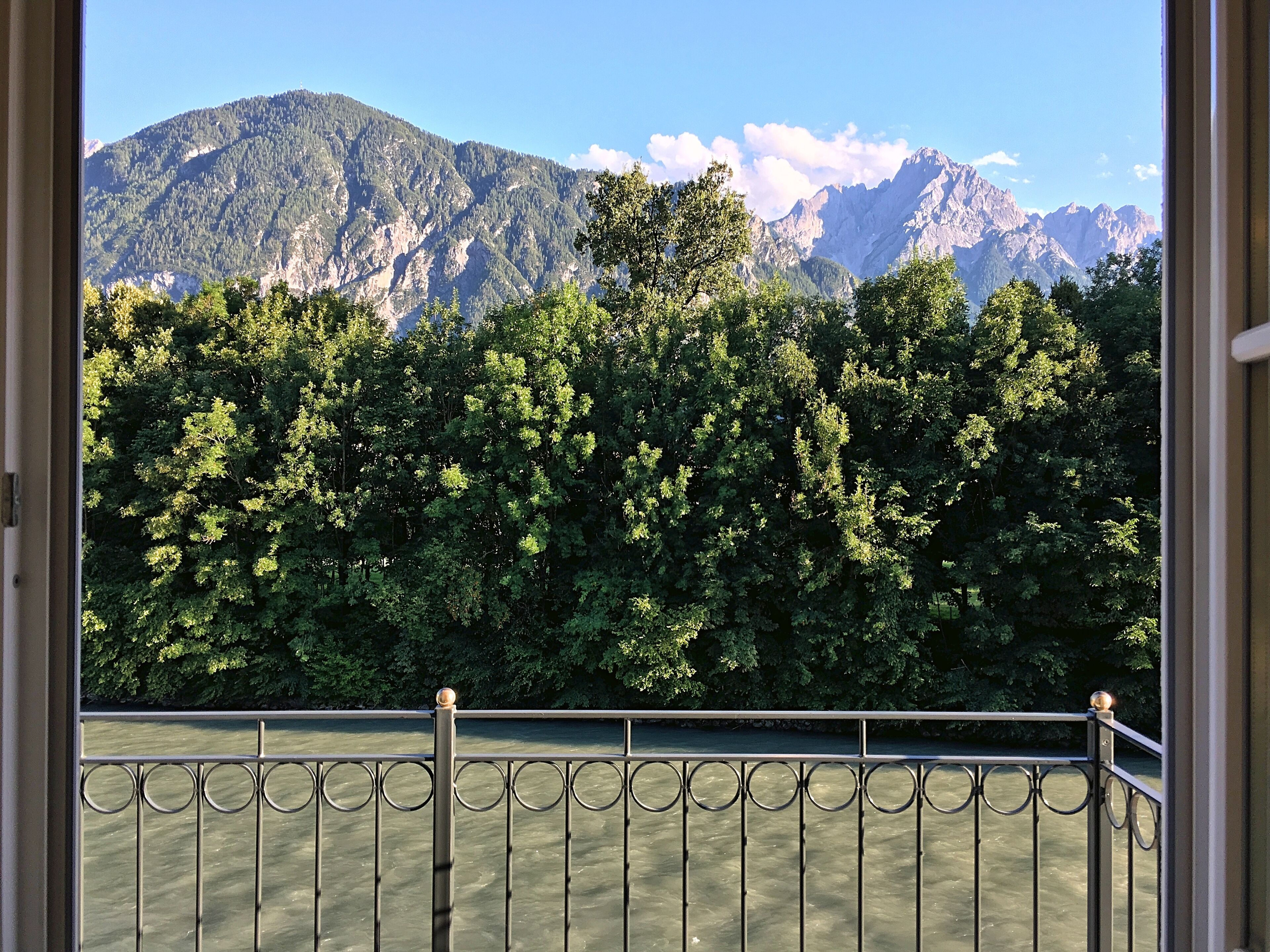 Vista de um rio com uma grade de metal em primeiro plano, uma floresta densa e montanhas ao fundo.