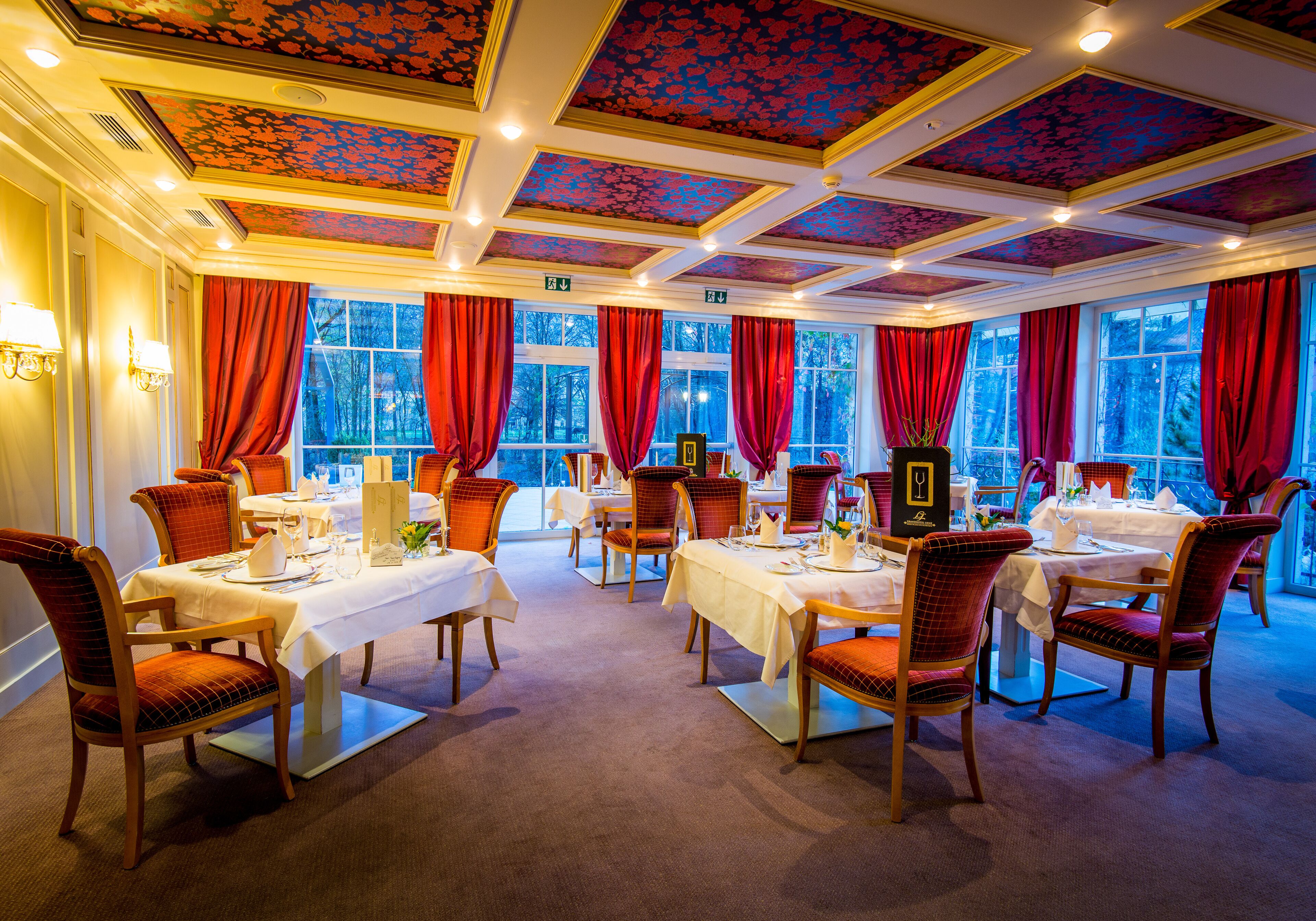 A dining area with multiple tables and chairs, large windows with red curtains, and a decorative ceiling.