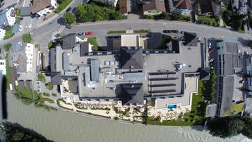 Eine Vogelperspektive auf einen Gebäudekomplex am Flussufer, bestehend aus mehreren Gebäuden, darunter ein großes, zentrales Gebäude mit geneigtem Dach, kleinere angrenzende Gebäude und einem Parkplatz.