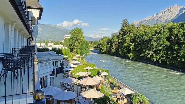 A riverside terrace with outdoor seating and a view of a mountain range. - Grandhotel Lienz (Lienz)