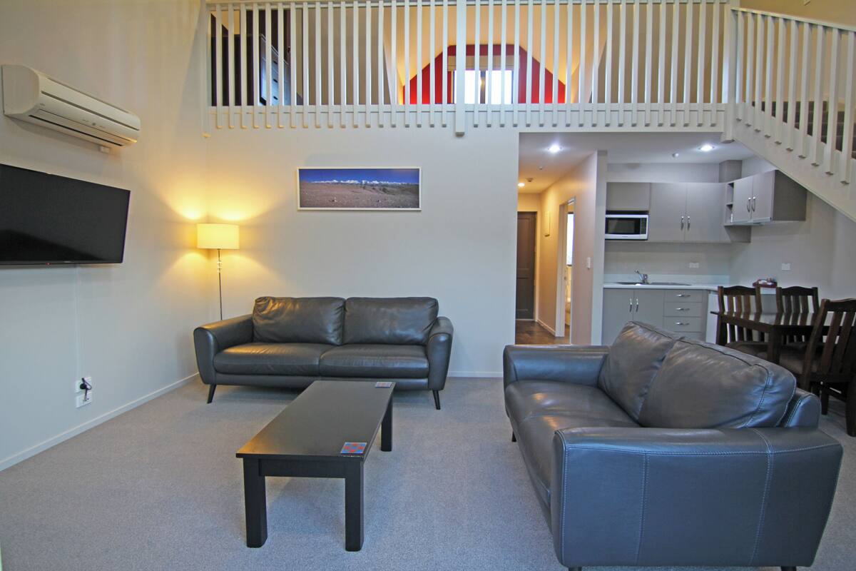 A living room with two leather sofas, a coffee table, a flat-screen TV, and a staircase leading to an upper level.