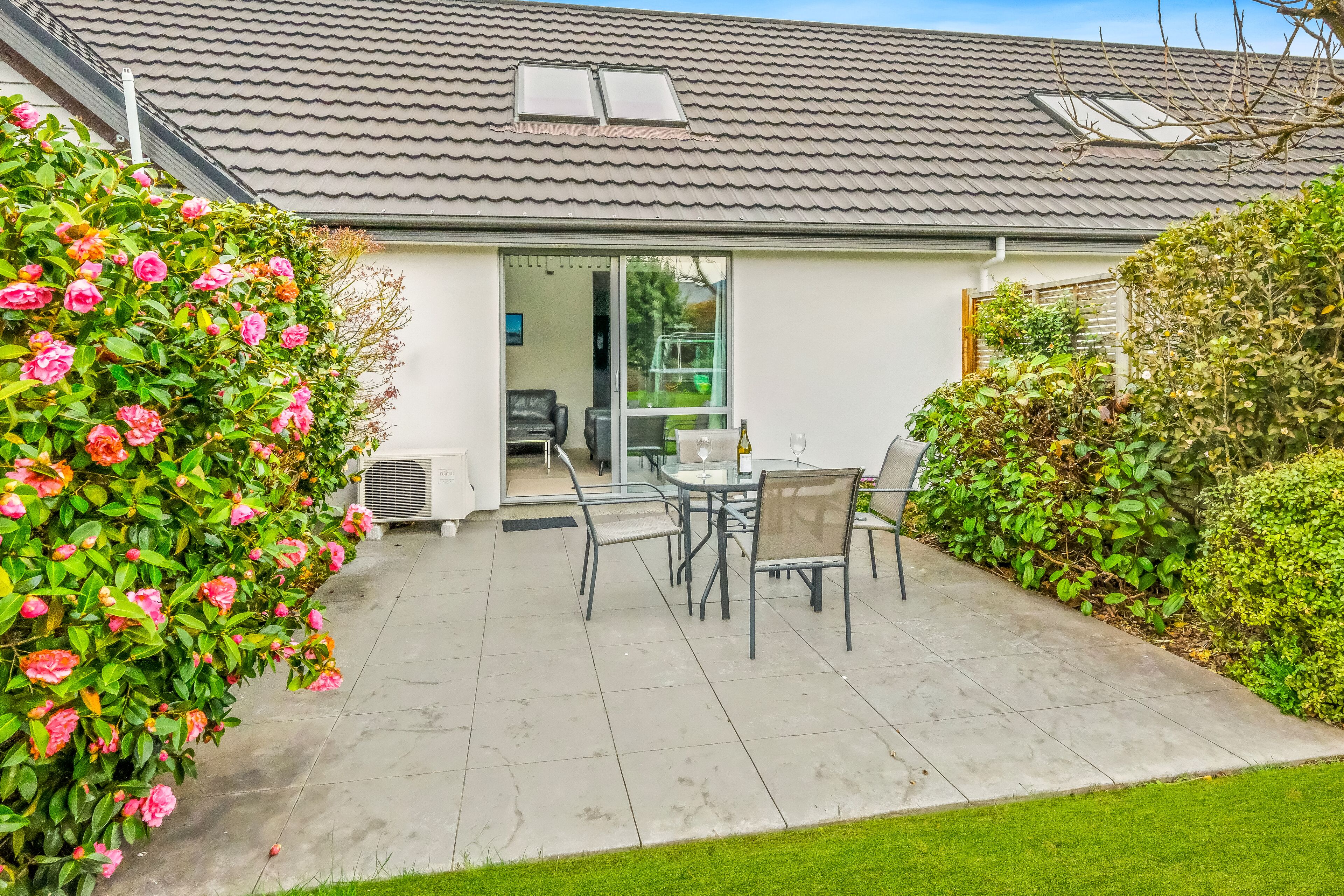 A patio with a table and chairs, a glass door leading to an interior space, and a garden with pink flowers.