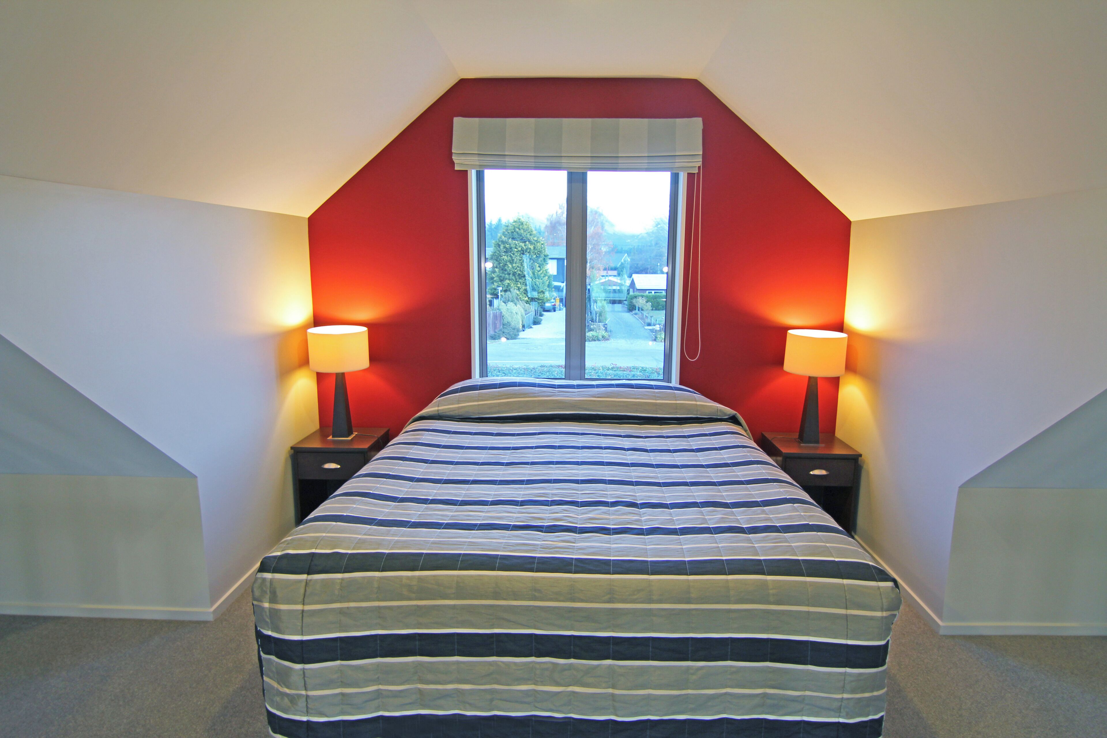 Un dormitorio con una cama grande, dos mesitas de noche con lámparas, una ventana con vistas al exterior y un techo inclinado.