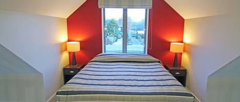 Un dormitorio con una cama grande, dos mesitas de noche con lámparas, una ventana con vistas al exterior y un techo inclinado.