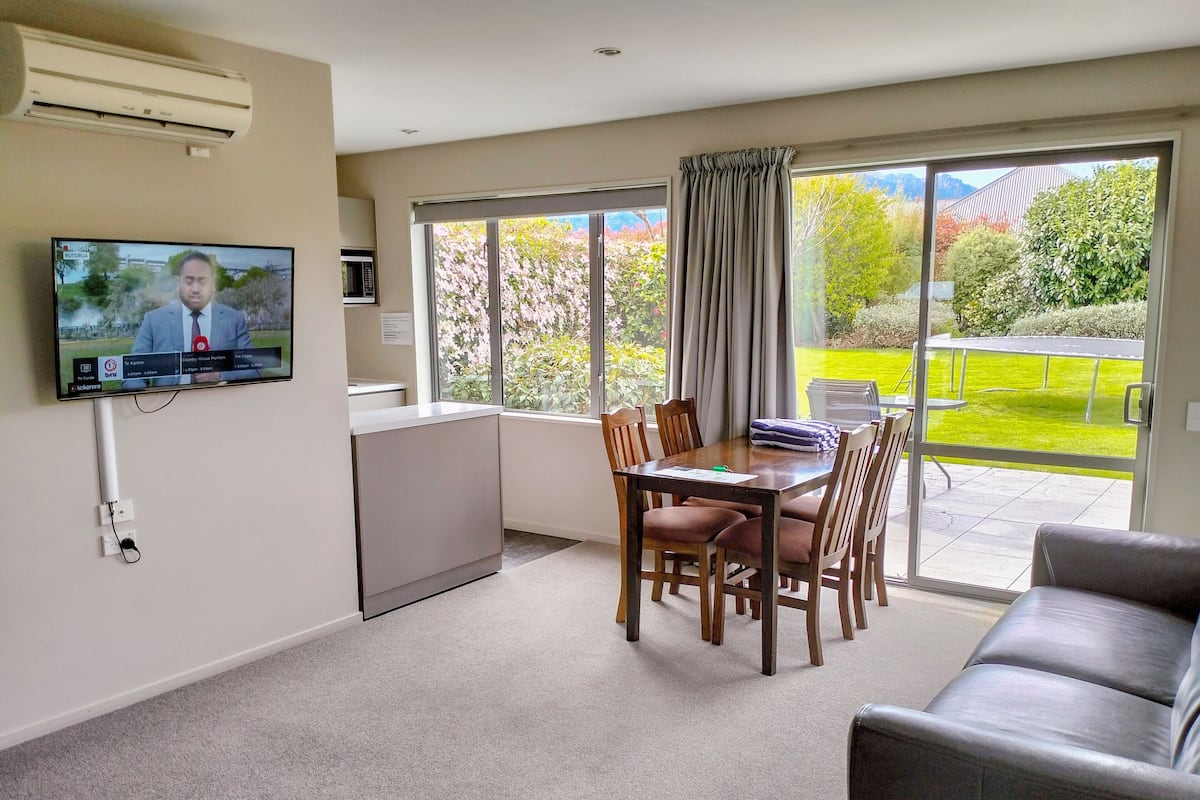 A modern living room with a flat-screen TV mounted on the wall, a dining area with a table and chairs, a sofa, and a sliding glass door leading to a balcony with a view of greenery and mountains.