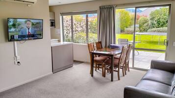 A modern living room with a flat-screen TV mounted on the wall, a dining area with a table and chairs, a sofa, and a sliding glass door leading to a balcony with a view of greenery and mountains.