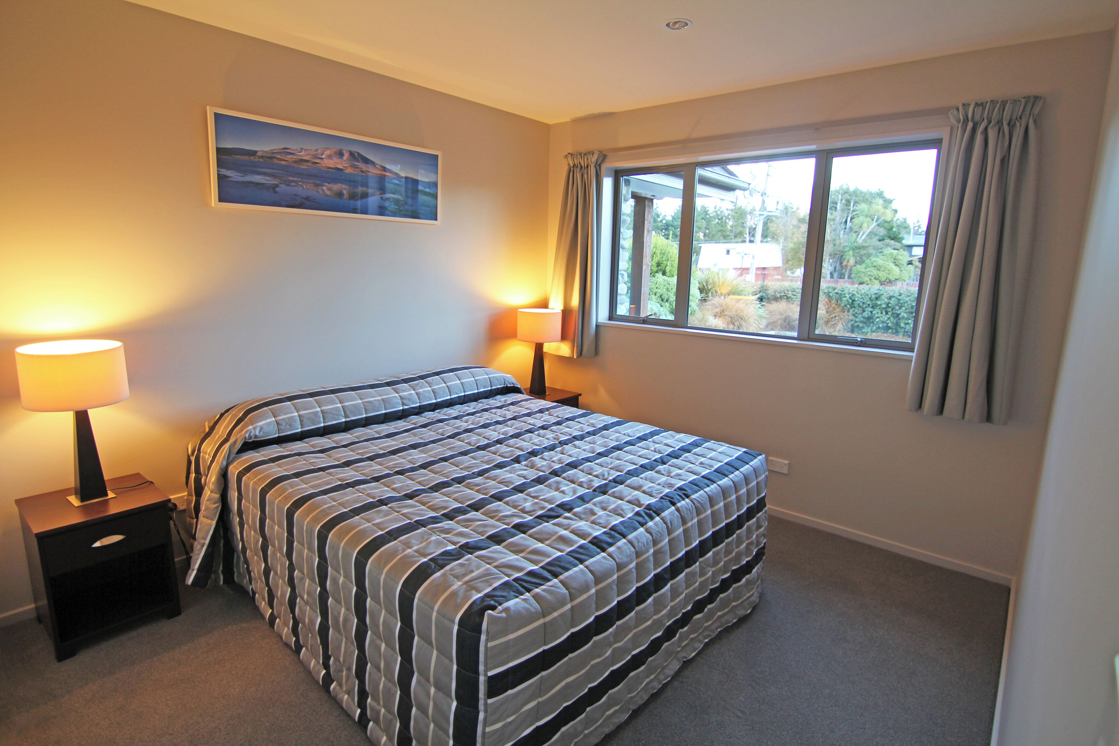 A bedroom with a bed, two bedside tables with lamps, a window with curtains, and a painting on the wall.