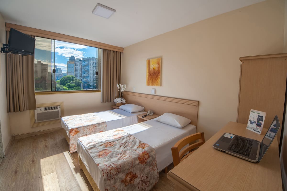 A hotel room with two beds, a desk with a laptop, a TV mounted on the wall, and a window with a city view.