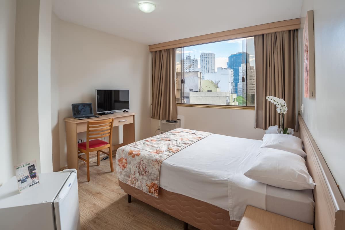 A hotel room with a bed, a desk with a TV, a chair, and a window with curtains.