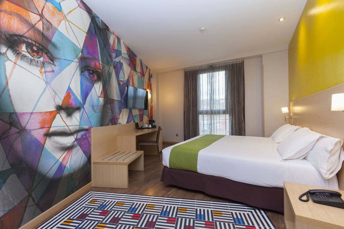 Una camera d'albergo con un letto grande, una scrivania, una sedia, una TV e un murale astratto dai colori vivaci.