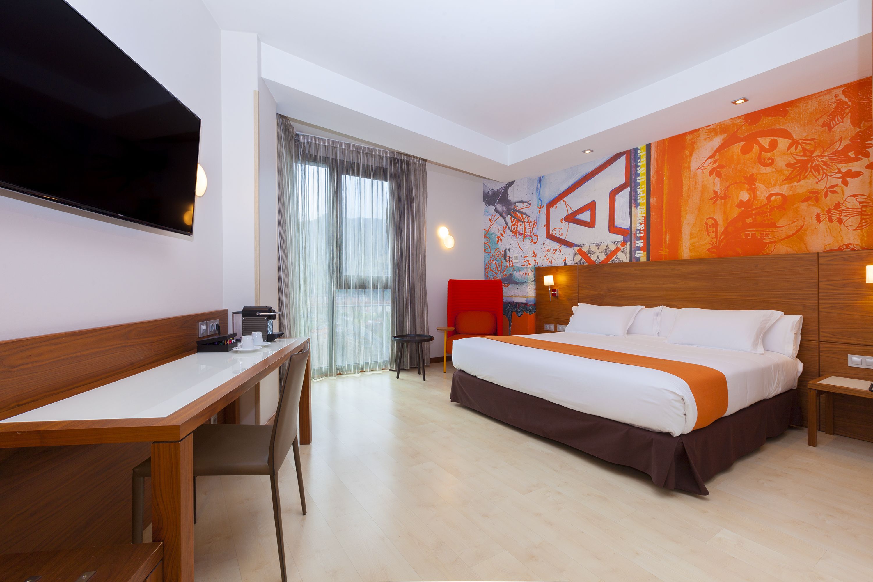 Una habitación de hotel con cama, escritorio, silla y televisor montado en la pared.