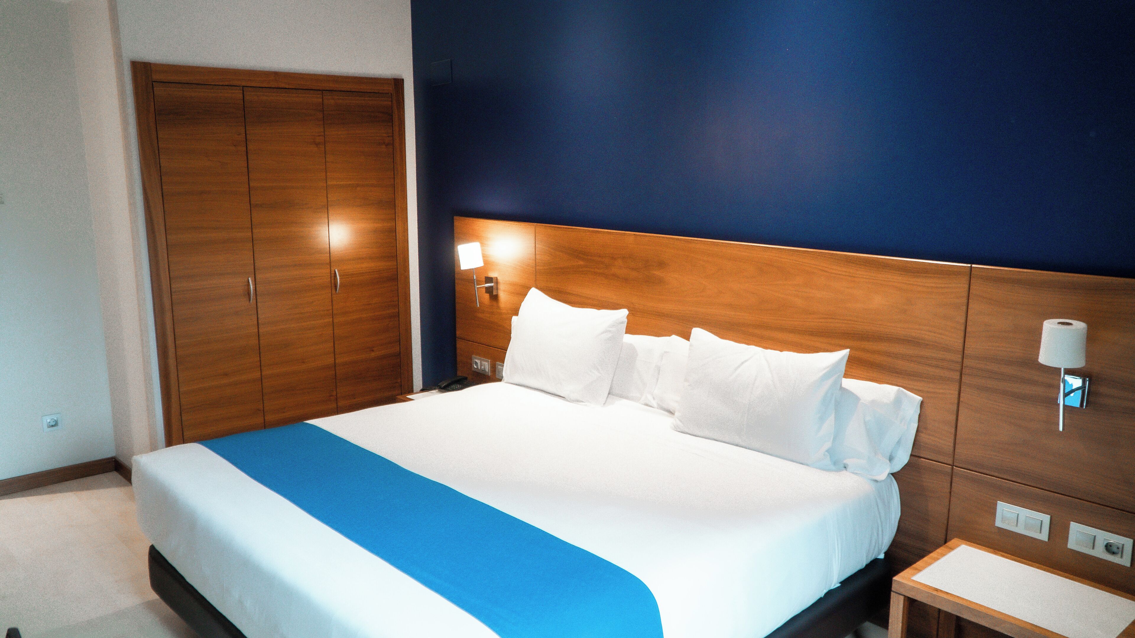 Una habitación de hotel con una cama grande, un armario de madera, lámparas de noche y una mesita pequeña.