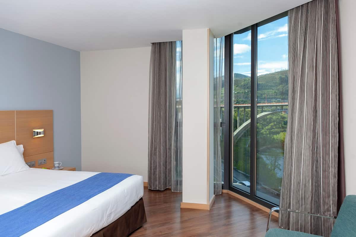 A hotel room with a large bed, a wooden headboard, a nightstand with a lamp, a view of a bridge and greenery through large windows, and a green chair.