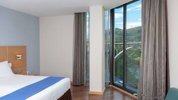 Habitación de hotel con una cama grande, cabecera de madera, mesita de noche con lámpara, vistas a un puente y follaje a través de amplias ventanas, y una silla verde.