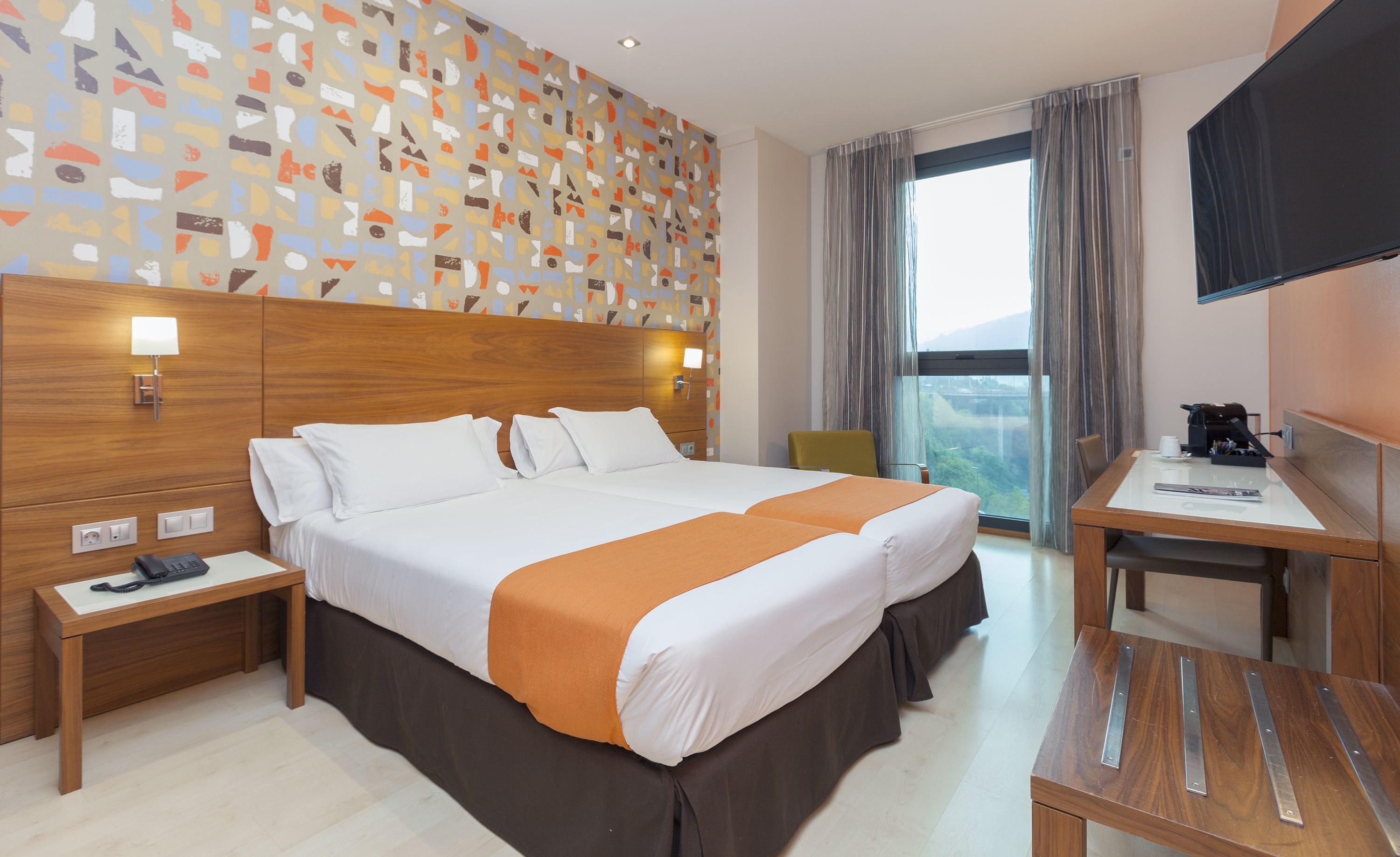 A hotel room with a large bed, a desk, a chair, a TV, and a window with a view.