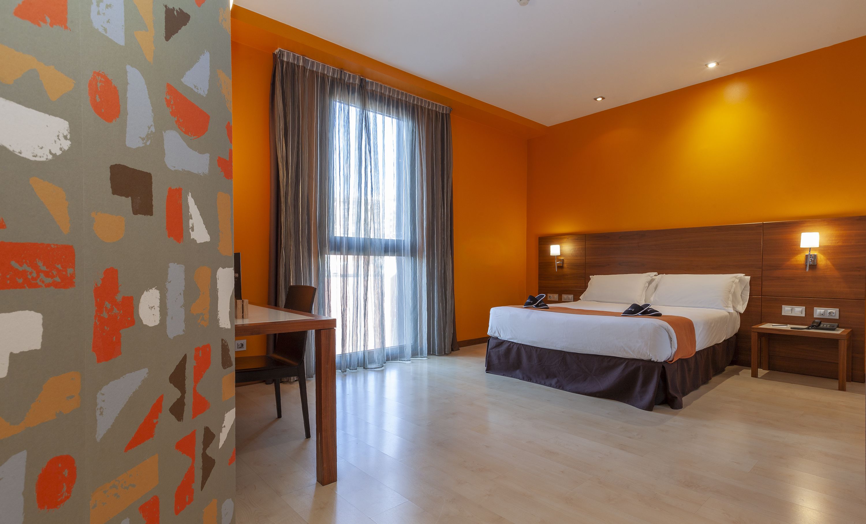 A hotel room with a bed, bedside tables, a desk, and a window with curtains.