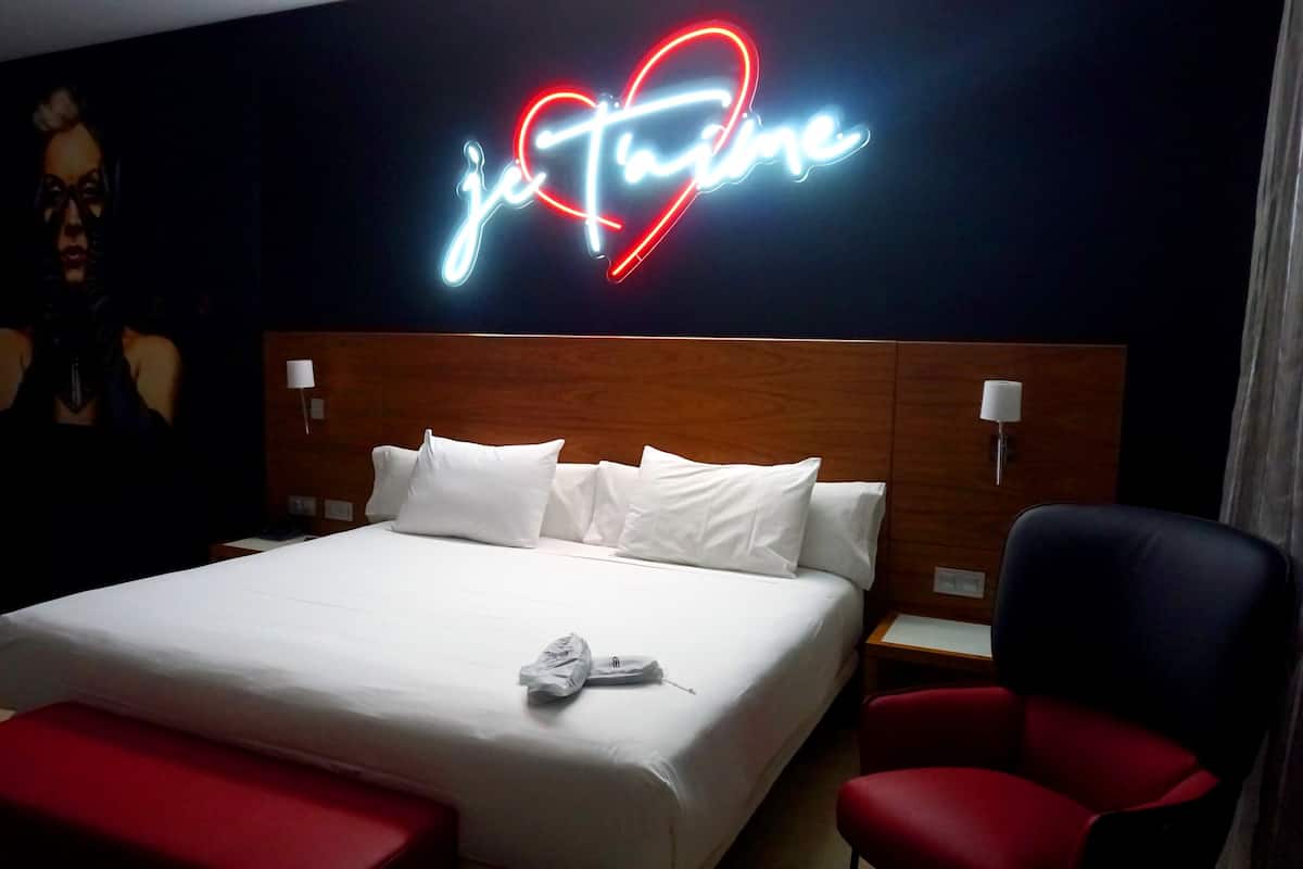 A hotel room with a large bed, white pillows, a red chair, a wooden headboard, and a neon sign that reads "je Time".