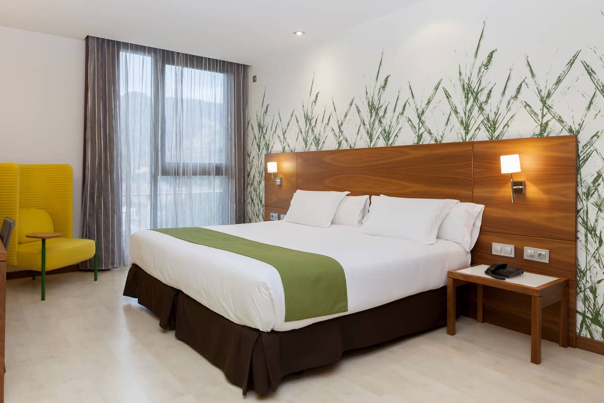 Una camera d'albergo con un letto grande, un comodino con un telefono, una poltrona gialla e una finestra con tende.