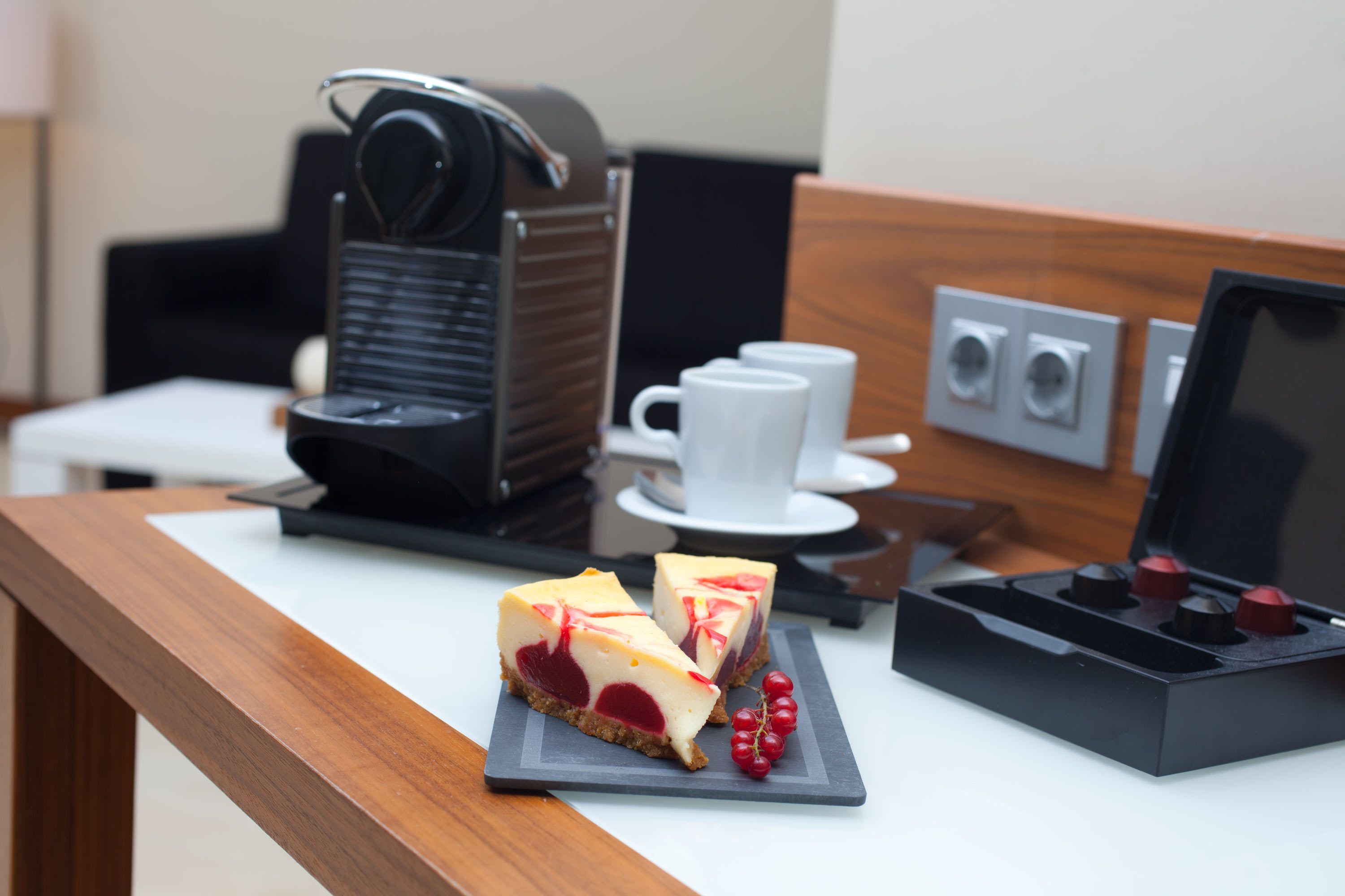 A slice of cheesecake on a slate plate with red currants, a coffee machine, a white cup, and a black box with red knobs on a hotel room table.