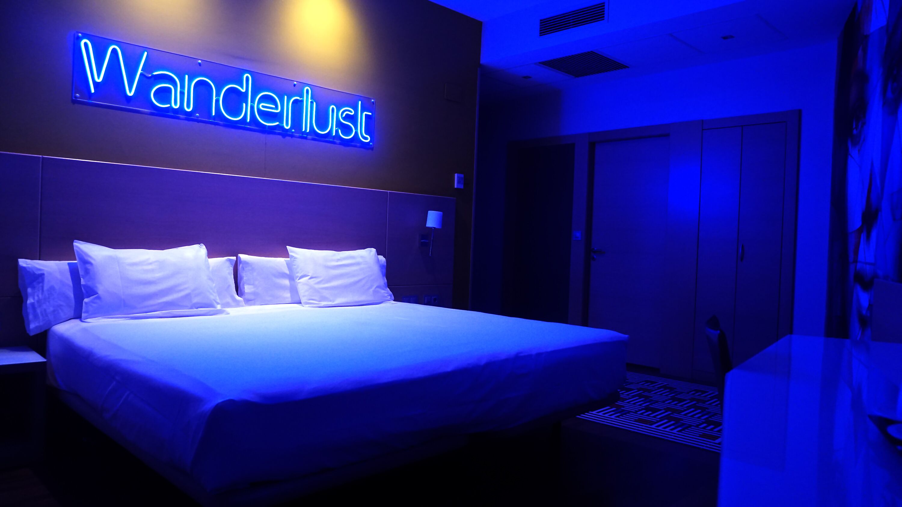Una camera d'albergo con un letto, un cartello al neon con la scritta “Wanderlust” e un armadio.