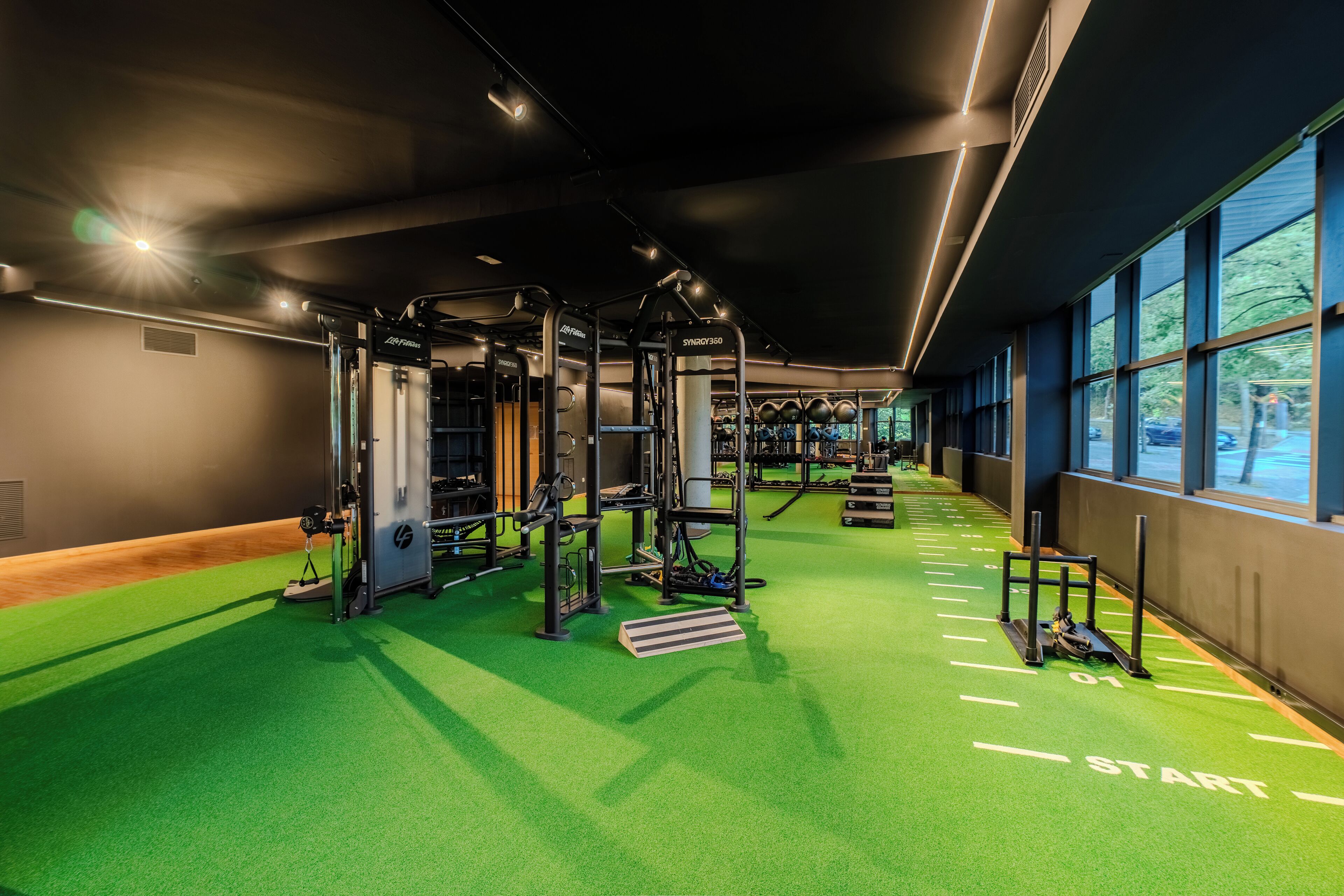 Un gimnasio con piso verde, equipamiento de ejercicios y amplias ventanas.