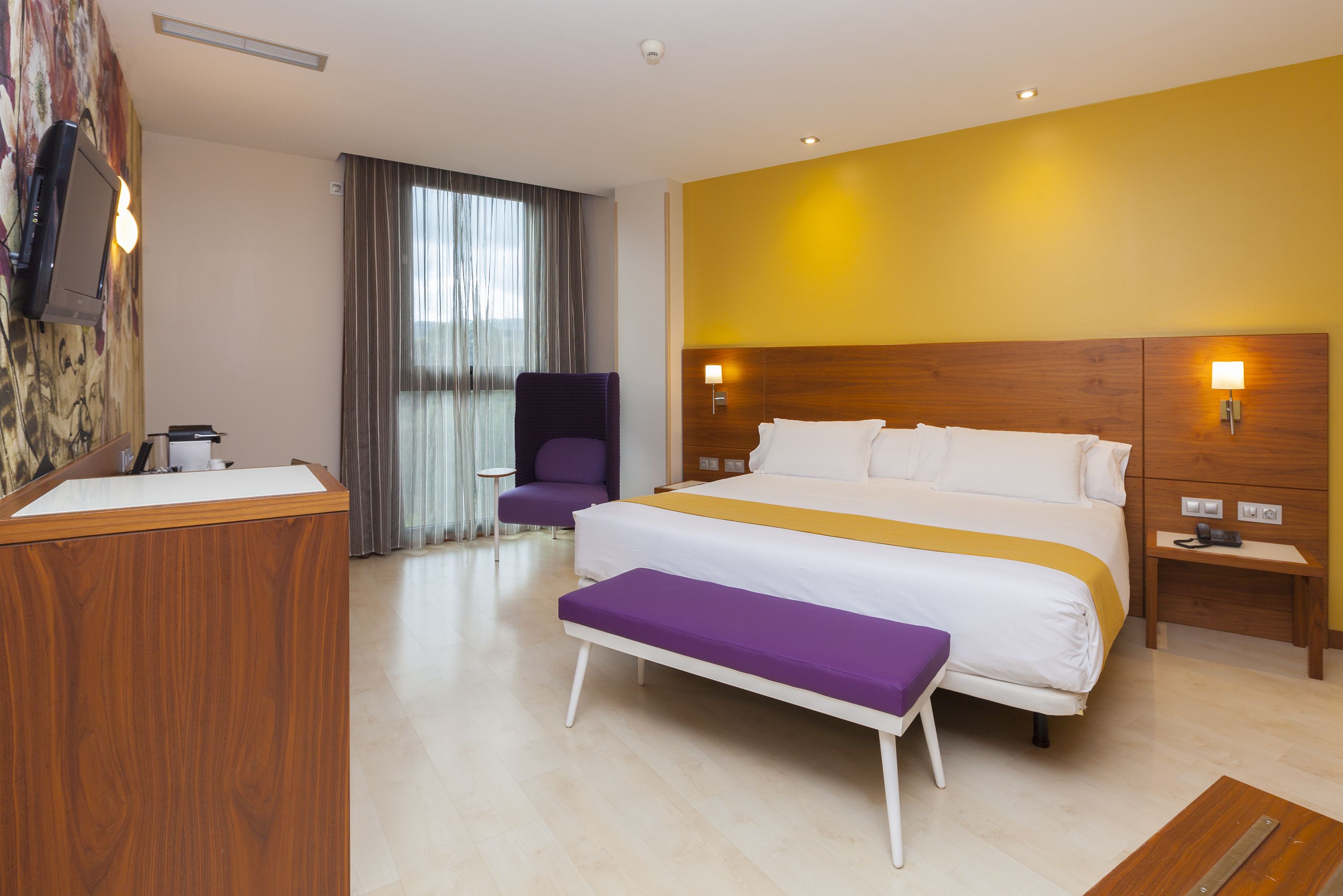 Habitación de hotel con una cama grande, un banco, un escritorio, una silla y un televisor instalado en la pared.